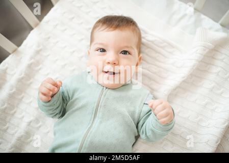 Funny smiling infant baby boy wake up in bed crib look at camera over white blanket top view closeup. Childhood. Good morning. Stock Photo