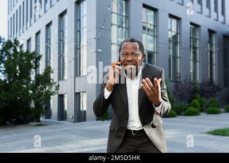 Angry african american boss arguing and shouting while talking on the phone, senior man in business suit outside office building. Stock Photo