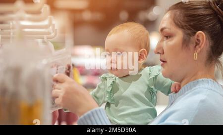 Baby girl with serious expression helps mother choose outfit for home. Stock Photo