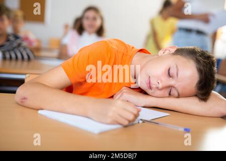 Bored tween schoolboy sleeping at desk in classroom during lesson Stock Photo