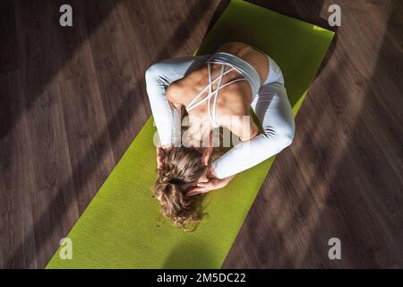 Young athletic woman leading a healthy lifestyle and practicing yoga performs kurmasana exercise, a variation of the turtle pose, trains while sitting Stock Photo