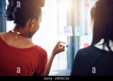 Every problem has a solution. Rearview shot of two businesswomen working on a glass wall the office. Stock Photo