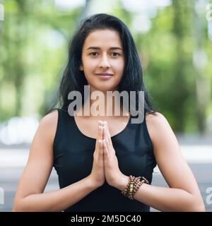 Portrait of young beautiful woman practicing yoga performing namaste greeting in park, instructor looking at camera Stock Photo