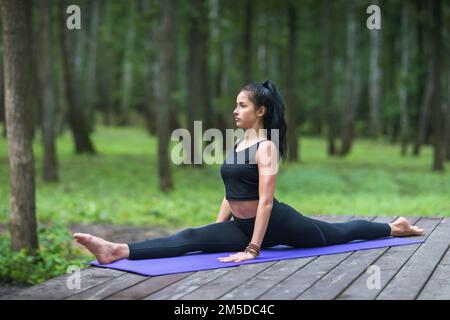 A young sports woman practicing yoga, doing a hanumanasana exercise, longitudinal split, trains out while sitting on a mat on a wooden platform in the Stock Photo