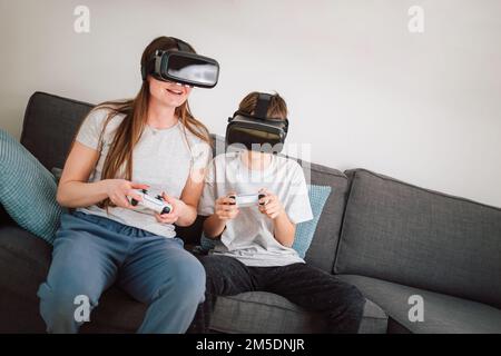 Mother and son enjoy entertainment video gaming together, using VR goggles and controllers while sitting on a sofa in the living room Stock Photo