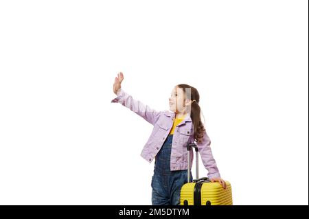 Adorable little girl traveler with yellow suitcase, waving hello with hand, looking at copy space on white background Stock Photo