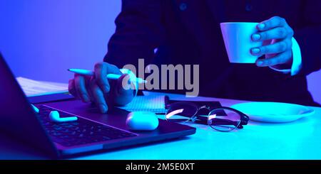 Making notes on laptop and drinking coffee. Working desk with computer, notebook, headphones and phone in neon colored light Stock Photo