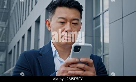 Bottom view pensive Asian Korean adult man middle-aged 40s businessman standing in city near office building looking up thinking browsing typing Stock Photo