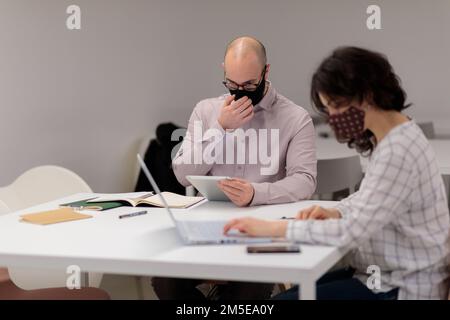Two young colleagues in masks sitting at a table, discussing and taking notes in the office Stock Photo