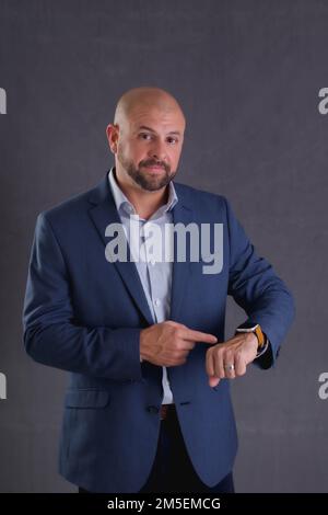 A Studio shot of a young bald businessman in a suit checking time standing against gray background Stock Photo