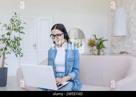 Beautiful young female student sitting on sofa at home with laptop and headset. He studies online, remotely. Stock Photo