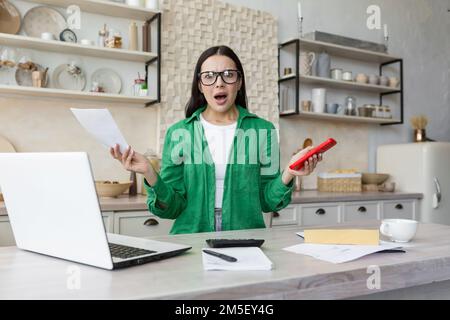 Portrait of worried and shocked woman sitting at home in kitchen with laptop and documents. Problems with accounts, credit, rent payment. Stock Photo