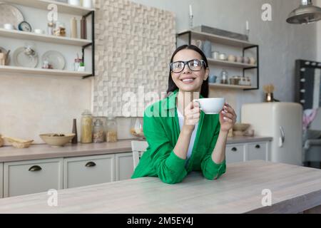 A young woman, businesswoman, freelancer, student drinks coffee and tea alone in the kitchen at home. Has a break. He holds a cup in his hands, rests, smiles, looks thoughtfully out the window. Stock Photo