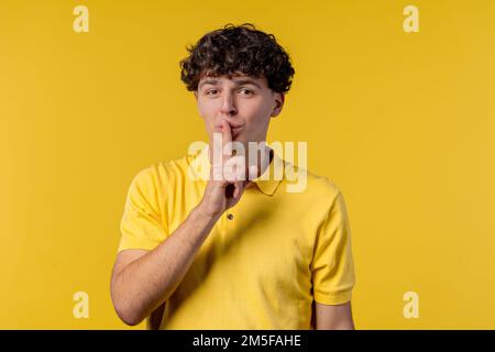 Handsome man with gesture of shhh, secret, silence, conspiracy, gossip concept. Smiling guy holding finger on lips, yellow studio background.  Stock Photo
