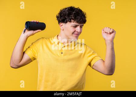 Man dancing, enjoying on yellow studio background. Guy moves to rhythm of music. Young teenager listening to music by wireless portable speaker - Stock Photo