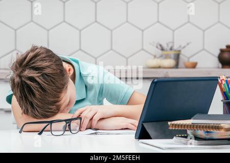 Portrait of young preteen weary boy pupil wearing blue T-shirt, lying sleeping on hand on desk near notebooks, glasses, color pencils, tablet at home Stock Photo