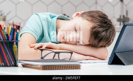 Portrait of young preteen tired boy wearing blue T-shirt, lying sleeping on hands on desk near notebooks, glasses, color pencils, tablet at home Stock Photo