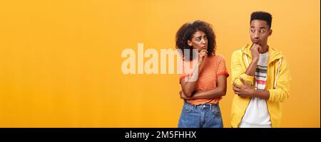 Silly confused girl standing gloomy and worried pursing lips and frowning holding hand on chin looking at boyfriend confused while guy glancing at Stock Photo