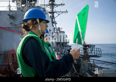 EAST CHINA SEA (March 16, 2022) Seaman Chelsea Lockwood, from Prescott, Arizona, signals to the Lewis and Clark-class dry cargo ship USNS Matthew Perry (T-AKE-9) during a replenishment-at-sea aboard the Arleigh Burke-class guided-missile destroyer USS Ralph Johnson (DDG 114). Ralph Johnson is assigned to Task Force 71/Destroyer Squadron (DESRON) 15, the Navy’s largest forward-deployed DESRON and the U.S. 7th fleet’s principal surface force. Stock Photo