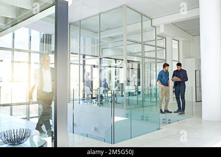 Another busy day in the office. two colleagues talking together over a digital tablet while standing in a modern office. Stock Photo
