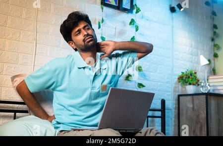 young man suffering from neck and back pain while using laptop on bed during work from home at night - concept of improper working posture, overworked Stock Photo