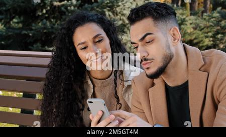 Hispanic ethnic couple sit on bench in autumn park man guy hold mobile phone browsing scrolling buying online with girl woman talk family smiling Stock Photo