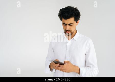 Studio portrait of handsome focused young man in shirt holding smartphone looking on device screen standing on white isolated background. Stock Photo