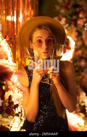 Funny portrait of girl in hat and pink glasses holding glasses of sparkling wine partying with blurred lights on background. Happy and fun emotions during night party. High quality vertical image Stock Photo