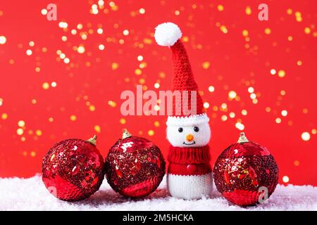 Knitted snowman in a red Christmas hat and sweater with three red balls on a red background with golden Christmas lights. Merry Christmas and New Year Stock Photo