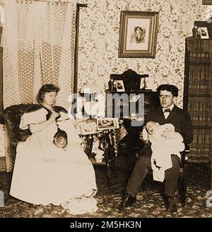 1897 - A domestic scene in a Victorian  Middle class household. Father nurses  the baby whilst mother does her craftwork, sowing or crocheting. Stock Photo