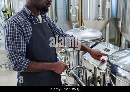 Close-up of African American worker in apron pushing the button to turn on the equipment for brewed beer Stock Photo