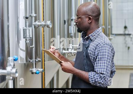 African American worker writing indicators of gauge in document while controlling the work at factory Stock Photo