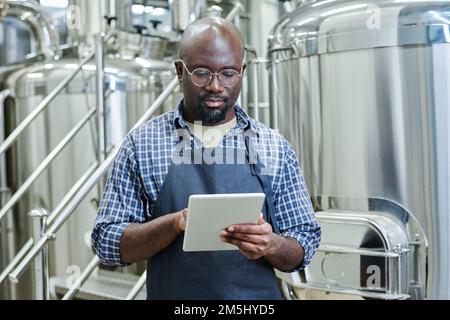 African American engineer concentrating on his online work using digital tablet during his work at brewery Stock Photo