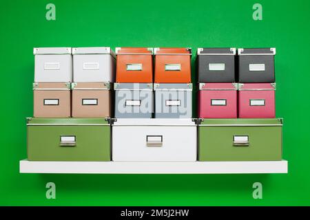 neat rows of boxes of different colors and sizes on a white bookshelf on a green wall. minimalistic composition on the theme of organization Stock Photo