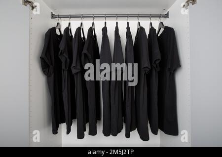 black t-shirts hang on a clothes rail in a white wardrobe. all t-shirts are the same color and size and are worn on hangers Stock Photo