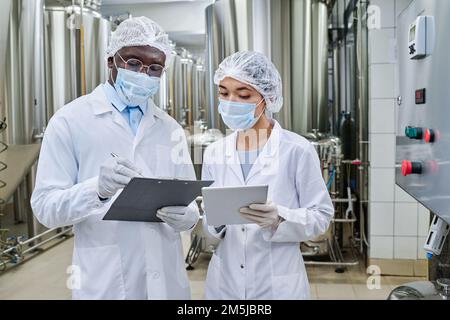 Engineers in masks and white coats working on tablet pc and making notes in document during their work at factory Stock Photo