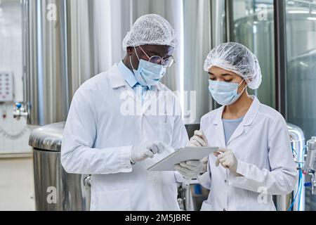 Two engineers in uniform and masks working on tablet pc together to control the equipment online Stock Photo