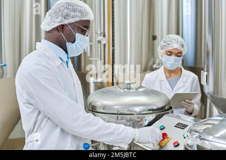 Engineer in protective clothing pushing button on equipment while working with assistant at factory Stock Photo