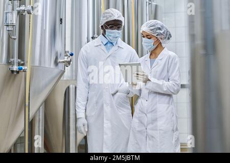 Two colleagues in uniform and protective masks discussing project on digital tablet during their work at brewery Stock Photo