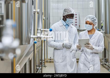 Two professional engineers in white coats and masks discussing something on digital tablet in workshop of brewery Stock Photo