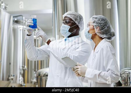 Multiethnic couple of engineers in uniform examining blue liquid in glass and making notes in tablet pc Stock Photo