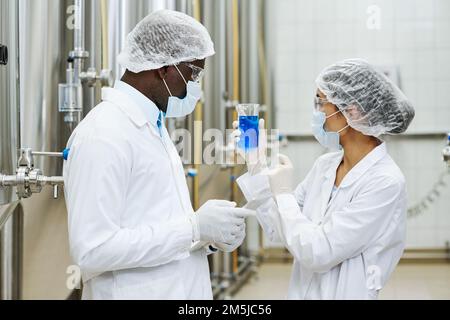 Scientists in uniform examining blue liquid in flask, they working in team and taking sample at factory Stock Photo