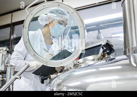 Female worker in protective wear writing equipment indicators in the card while working with machinery at factory Stock Photo