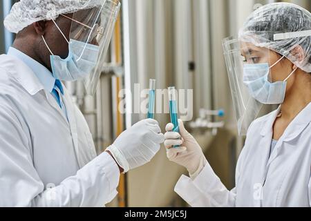 Two scientists in protective mask hoding test tubes with blue liquid in their hands while conducting experiment at plant Stock Photo