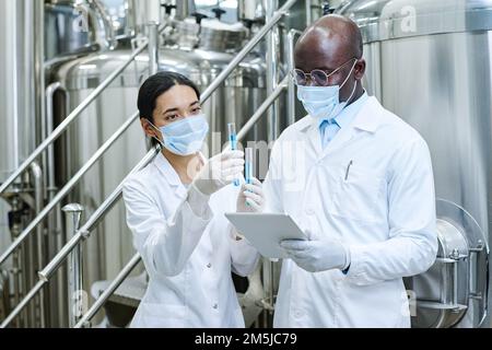 Couple of scientists in white coats examining liquid in test tubes and checking results using digital tablet standing in workshop Stock Photo