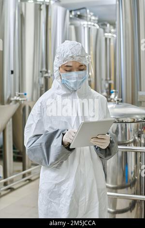 Young engineer in protective wear and mask working on tablet pc standing in workshop Stock Photo