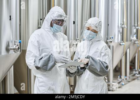Workers in protective wear discussing online project on digital tablet during teamwork at chemical factory Stock Photo