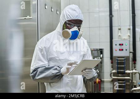 African American engineer in protective workwear and mask using digital tablet, he working in harmful production Stock Photo