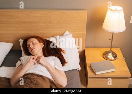 Portrait of sick tired young woman with flu lying under blanket with closed eyes on bed holding thermometer in armpit. Stock Photo