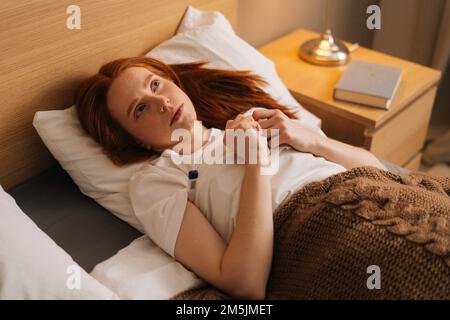 High-angle view of sick young woman with flu lying under blanket on bed holding thermometer in armpit. Ill female with cold lying in bed with high Stock Photo